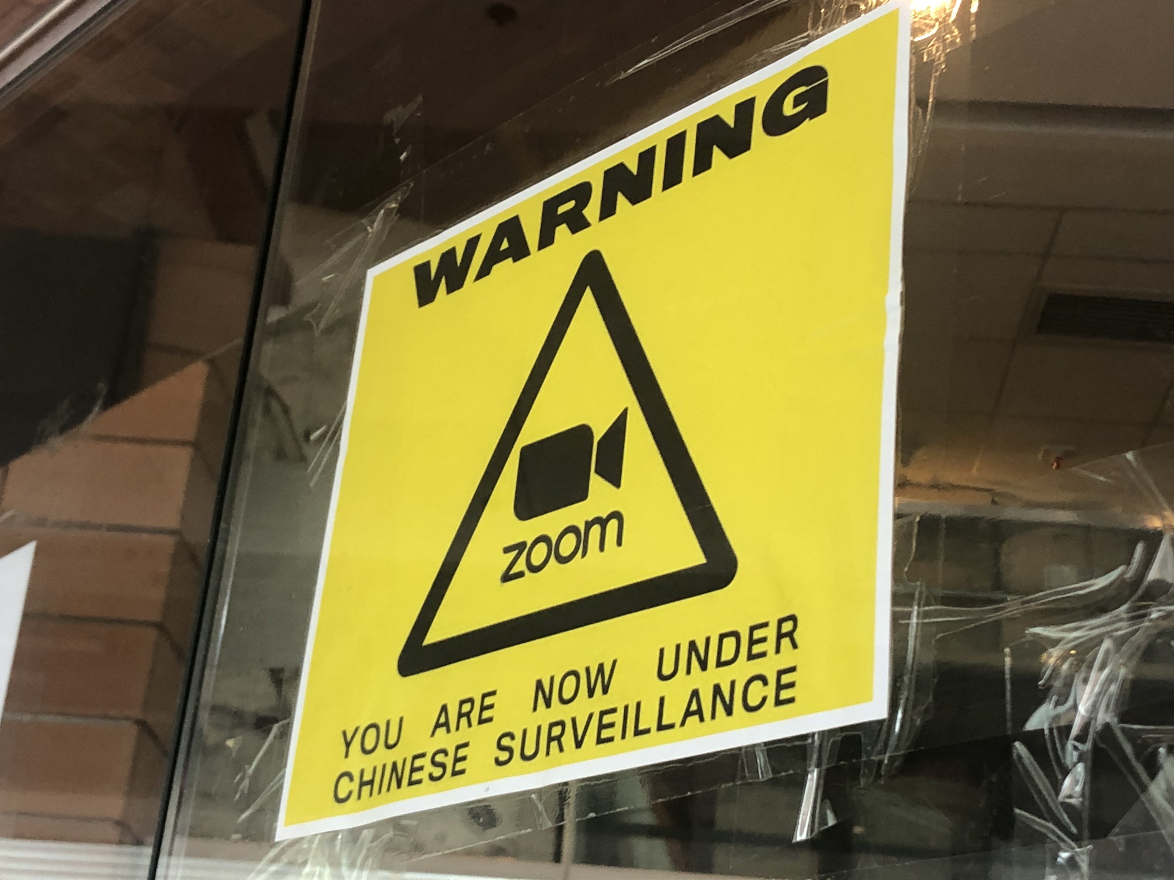 Fig. 2. Personal photograph of an anti-Zoom poster, equating Zoom with “surveillance,” appended to a campus window. This photograph depicts a square yellow sign on a window. The sign contains the word “WARNING” at the top in black text. Beneath is a black triangle with the Zoom logo (a video camera and the word “zoom” in lowercase font) inside. Under the triangle is the phrase “YOU ARE NOW UNDER CHINESE SURVEILLANCE.”