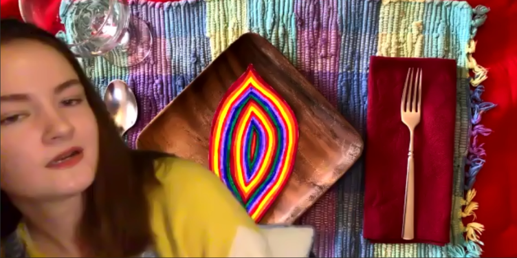 Fig. 12. Luisa Maria Lynch, Pacita Abad Virtual Presentation with Place Setting. This image depicts a Zoom window in which a person in a yellow shirt sits in front of a virtual place setting. The place setting contains a wooden plate with a multicolored design. On the left of the plate is a silver spoon, and on the right is a silver fork atop a red napkin. These objects rest on a multicolored woven placemat.