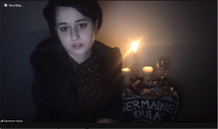 Fig. 13. Sarah Boline, Germaine Dulac Virtual Presentation with Place Setting. This image depicts a Zoom window in which a student, dressed in black, sits in a dark room in front of a table with candles, a clear wine glass, and other items. In front of the table is a banner reading “Germaine Dulac.”